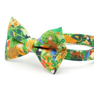 May include: A green cat collar with a bow tie featuring a jungle print with toucans, monkeys, tigers, and other animals. The collar has a gold bell.