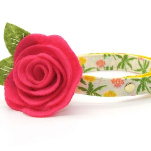 May include: A pet collar featuring a bright pink felt rose with two green felt leaves attached to the side. The collar band has a white background with a pattern of green palm trees and small yellow and orange flowers, finished with a gold bell.