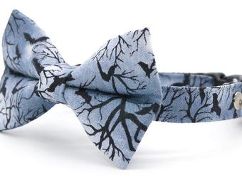 May include: A light blue pet collar featuring a bow tie with a black silhouette pattern of bare tree branches and flying birds. The collar includes a silver-toned metal ring and a small, round silver-toned bell attached to the side of the strap.
