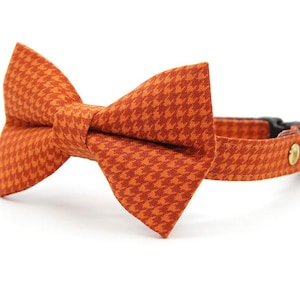 May include: An orange and dark orange houndstooth patterned pet collar featuring a matching fabric bow tie attached to the front. The collar includes a black plastic buckle, a gold-toned metal rivet, and a small round gold-toned metal bell.