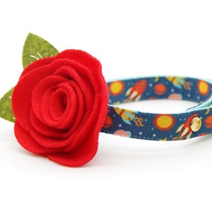 May include: A red felt rose flower attached to a blue cat collar with a rocket ship and planet pattern. The collar has a gold bell.
