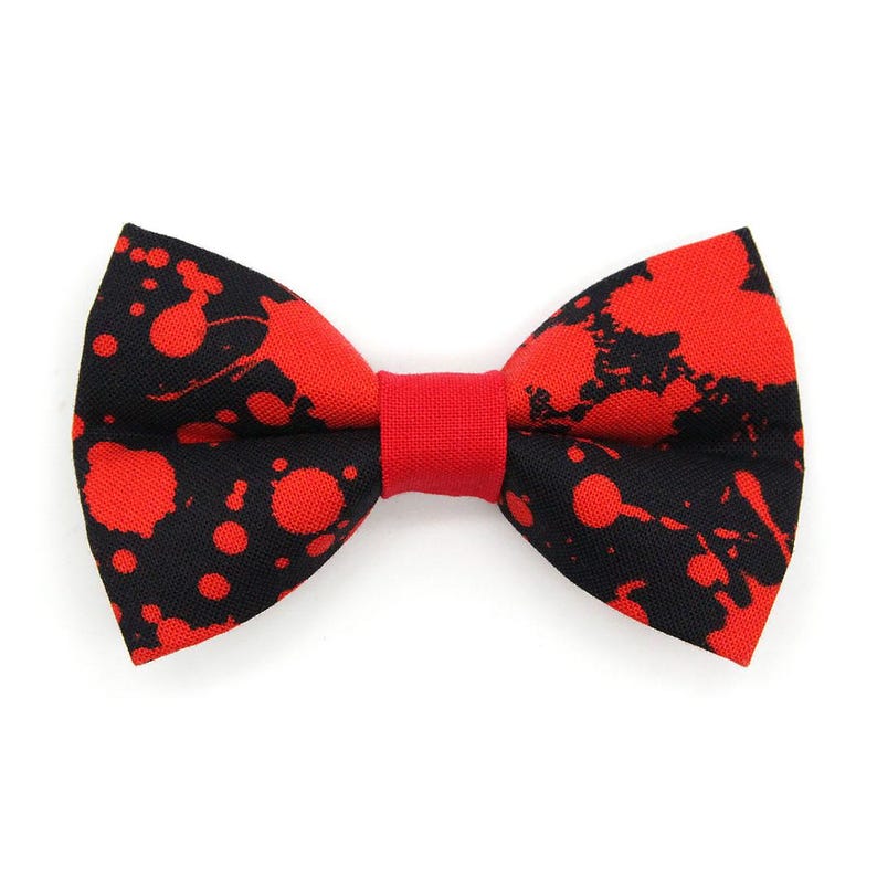 May include: A bow tie featuring a black fabric base with a vibrant red paint splatter pattern. The center of the bow tie is cinched with a solid red fabric band, creating a distinct contrast against the dark background of the accessory.