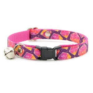 May include: A pink cat collar with a bell and a black buckle. The collar is decorated with a pattern of dice with a purple, orange, and yellow color scheme.