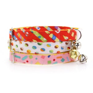 May include: Three colorful cat collars with bell charms. The collars are made of fabric with different patterns: red with yellow stripes, white with colorful polka dots, and pink with yellow confetti.