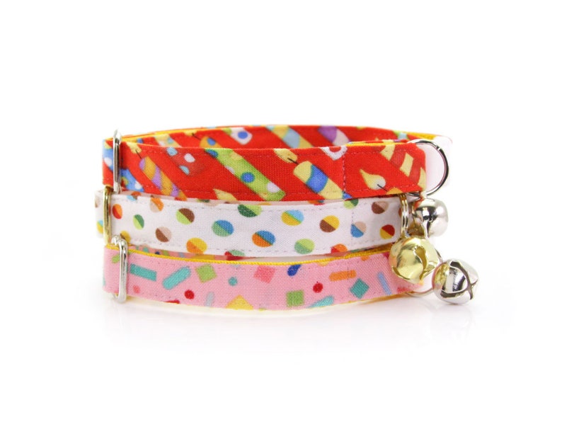May include: Three colorful cat collars with bell charms. The collars are made of fabric with different patterns: red with yellow stripes, white with colorful polka dots, and pink with yellow confetti.