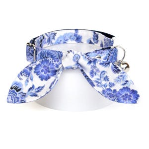 May include: Blue and white floral cat collar with a bell. The collar is made of fabric and has a bow tie design.