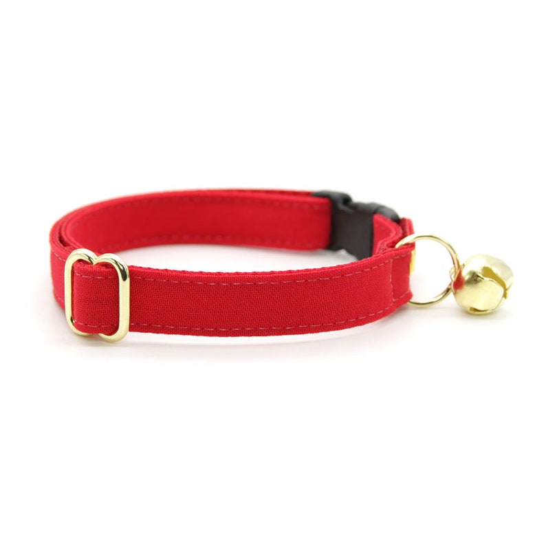 May include: A red cat collar with a gold buckle and a gold bell. The collar is made of a soft, comfortable fabric.