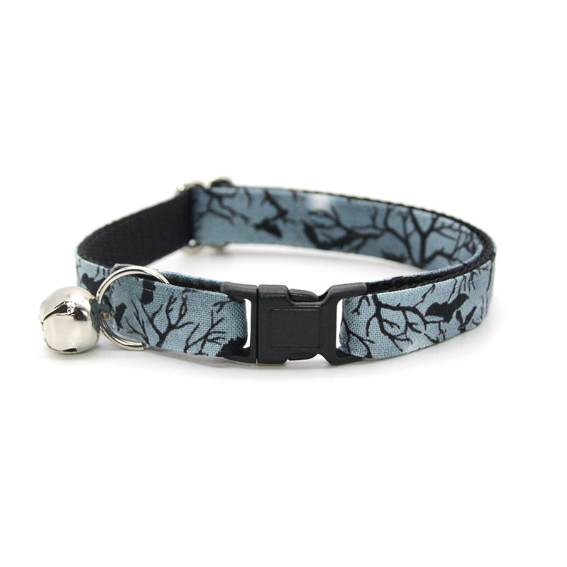 May include: A blue and black cat collar with a breakaway buckle and a silver bell. The collar features a black and white pattern of branches and birds.