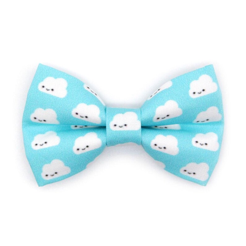 May include: A light blue bow tie with a repeating pattern of smiling white cloud shapes. The bow tie is tied in the center and has a classic bow shape. The background is white.