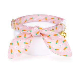 May include: Pink cat collar with a bow tie featuring a white bunny and carrot pattern. The collar has a gold bell.