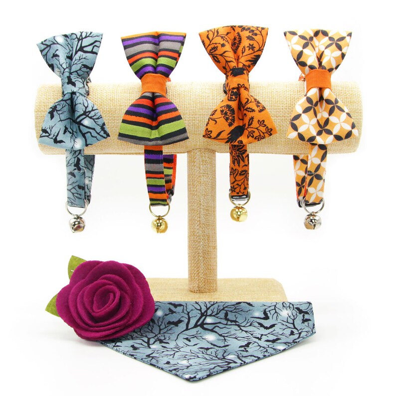 May include: Four different cat collars with bow ties on a display stand. The collars are made of fabric with various patterns, including a spooky tree design, a striped design, a floral design, and a geometric design. Each collar has a bell attached.