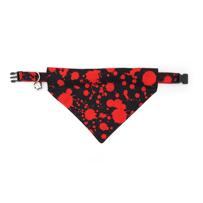 May include: A black pet collar with an attached triangular bandana featuring a red paint splatter pattern. The collar includes a black plastic side-release buckle and a small silver-toned metal bell attached to a metal D-ring for secure fastening.