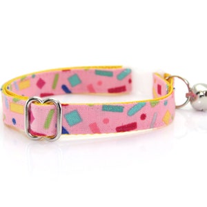 May include: A pink cat collar with a yellow border and a colorful confetti pattern. The collar has a silver buckle and a silver bell.