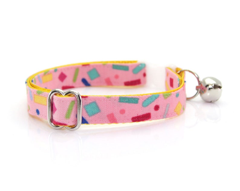 May include: A pink cat collar with a yellow border and a colorful confetti pattern. The collar has a silver buckle and a silver bell.