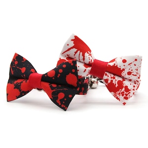 Op de afbeelding: Twee strikjes voor huisdieren met een rood verfspattenpatroon. E&eacute;n strikje heeft een zwarte achtergrond met rode spatten, de andere een witte achtergrond met rode spatten. Elk strikje heeft een effen rode middenband en een klein metalen belletje.