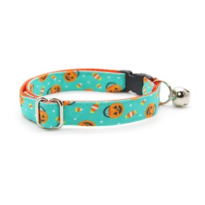 May include: A turquoise blue cat collar with a Halloween themed print of orange jack-o'-lanterns and candy corn. The collar has a silver buckle and a silver bell.