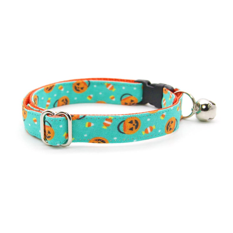 May include: A turquoise blue cat collar with a Halloween themed print of orange jack-o'-lanterns and candy corn. The collar has a silver buckle and a silver bell.