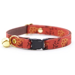May include: A red cat collar with a floral pattern in yellow and brown. The collar has a black plastic buckle, a gold-colored bell, and gold-colored metal hardware. The collar is approximately 0.5 inches wide.