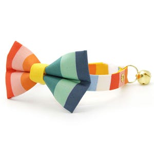 May include: A pet collar featuring a bow tie with a multi-colored striped pattern in shades of orange, pink, yellow, green, and blue. The collar includes a gold-toned metal bell attached to a ring, with a yellow fabric center on the bow tie.