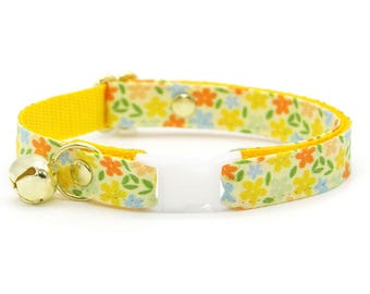 May include: A yellow cat collar with a floral pattern in orange, yellow, blue, and green. The collar has a white plastic buckle, a gold-colored bell, and gold-colored metal hardware.