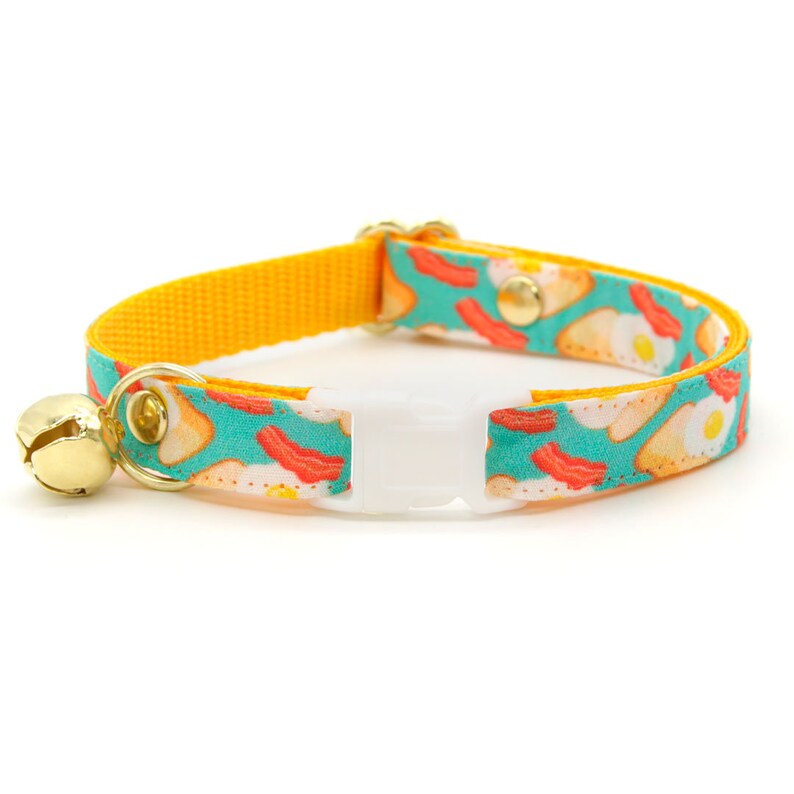 K&ouml;nnte beinhalten: Ein gelbes und t&uuml;rkisfarbenes Katzenhalsband mit einem Fr&uuml;hst&uuml;cks-Motiv mit Eiern und Speck. Das Halsband hat eine wei&szlig;e Kunststoffschnalle und eine goldene Glocke.