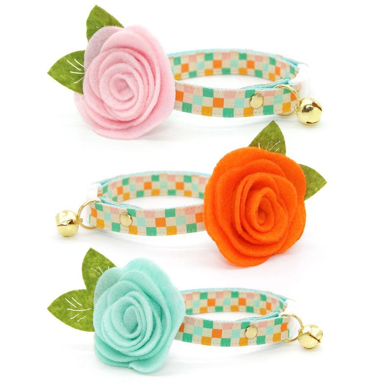 K&ouml;nnte beinhalten: Drei Katzenhalsb&auml;nder mit floralen Akzenten. Jedes Halsband hat eine andere farbige Filzblume: rosa, orange und t&uuml;rkis. Die Halsb&auml;nder haben ein kariertes Muster in Pastellfarben und eine kleine goldene Glocke.
