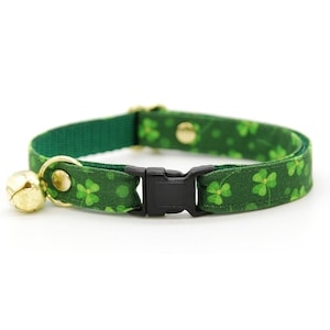 May include: Green cat collar with a shamrock pattern and a gold-colored bell. The collar has a black plastic buckle and a gold-colored metal clasp. The collar is approximately 12 inches in length.