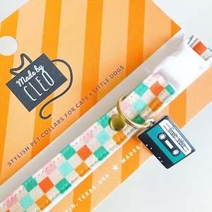 K&ouml;nnte beinhalten: Ein Haustierhalsband mit einem farbenfrohen Karomuster in Rosa, Orange und T&uuml;rkis. Das Halsband hat einen goldfarbenen Ring und einen schwarzen Kassettenband-Anh&auml;nger mit der Aufschrift "Good Vibes". Die Verpackung tr&auml;gt die Aufschrift "Made by CLEO".