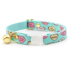 May include: A turquoise blue cat collar with a bell and a white plastic buckle. The collar is decorated with a repeating pattern of pink and yellow candy hearts with various messages, including "Be Mine", "True Love", "Hugs", and "My BFF".