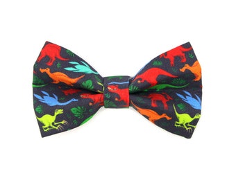 May include: A black bow tie with a colorful dinosaur print. The dinosaurs are in shades of red, orange, yellow, green, and blue.