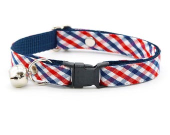 May include: A pet collar featuring a red, white, and blue diagonal gingham check pattern. The collar includes a black plastic side-release buckle, a silver-toned metal bell attached to a split ring, and a silver-toned metal adjustment slider.