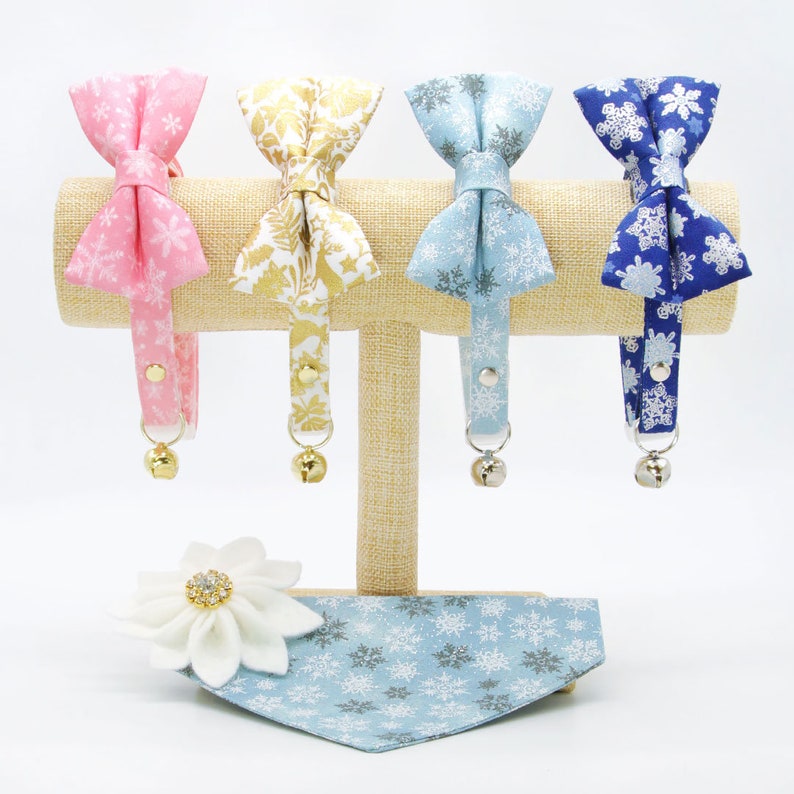 May include: Four cat collars with different patterned bow ties and bells. The collars are displayed on a wooden stand. A blue bandana with a snowflake pattern is also shown.