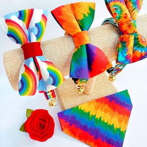 Może przedstawiać: Trzy kolorowe obroże dla psa w stylu tie-dye z wzorami tęczy. Obroże są prezentowane na brązowym stojaku. Jedna obroża ma wz&oacute;r tęczy i chmur, druga ma wz&oacute;r tie-dye, a trzecia ma wz&oacute;r tęczy i chmur.
