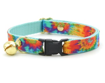 Puede incluir: Un collar para gato azul turquesa con un patrón tie-dye en tonos naranja, amarillo, rojo y morado. El collar tiene una hebilla de plástico negro y una campanilla dorada.