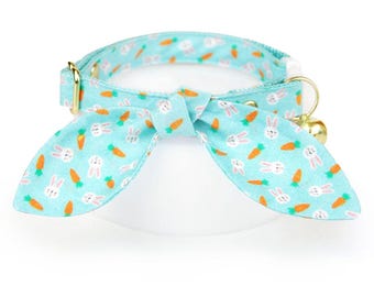 May include: A light blue cat collar with a bunny and carrot pattern and a gold bell. The collar has a bow tie shape.