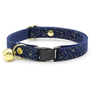 Könnte beinhalten: Ein marineblaues Katzenhalsband mit goldfarbenem Glitzermuster und einer goldenen Glocke. Das Halsband hat eine schwarze Kunststoffschnalle.