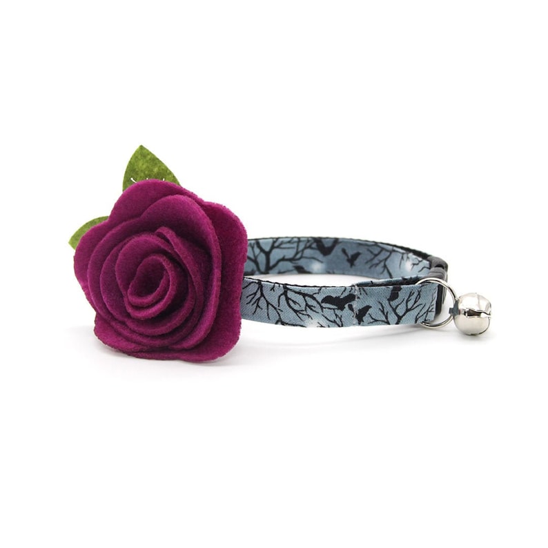 May include: A purple felt flower with green leaves attached to a blue and black cat collar with a silver bell.