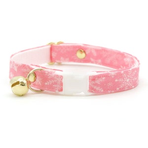 May include: Pink cat collar with a white snowflake pattern and a gold bell. The collar has a white plastic buckle.
