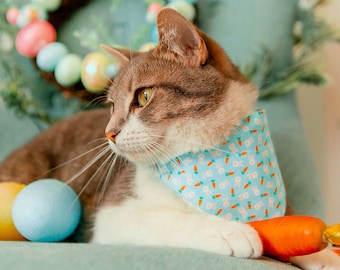 May include: A gray and white cat wearing a light blue bandana with a white bunny and carrot pattern. The cat is lying on a light blue surface with Easter eggs in the background.