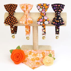 Pu&ograve; includere: Quattro collari per gatti con diversi motivi, tra cui zucche arancioni e rosa, a scacchi neri e arancioni e stelle nere con stelle rosa e arancioni. I collari sono esposti su un supporto in legno con un bandana floreale arancione e rosa abbinato e due fiori di feltro.