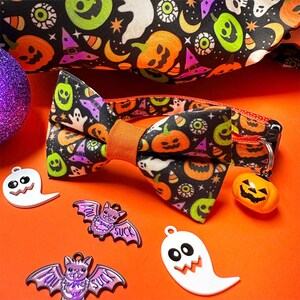 Puede incluir: Un collar para mascotas con tem&aacute;tica de Halloween con una pajarita estampada con calabazas, fantasmas y sombreros de bruja. El collar tiene un dije de calabaza y est&aacute; rodeado de dijes con tem&aacute;tica de Halloween, todo sobre una superficie naranja.