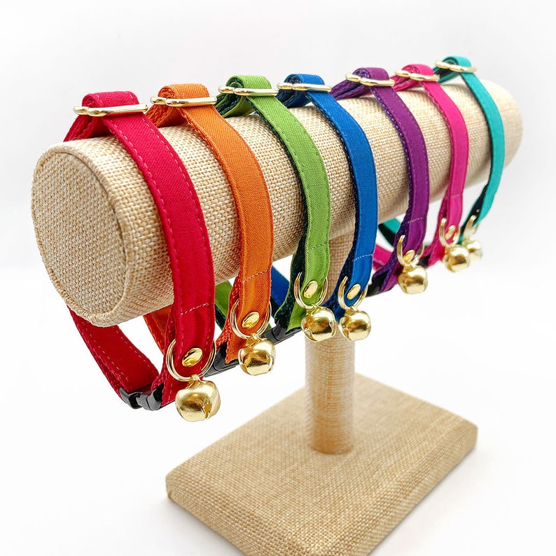 May include: A set of colorful cat collars with bells. The collars are displayed on a burlap stand and come in a variety of colors, including red, orange, green, blue, purple, and pink.