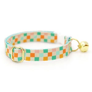 K&ouml;nnte beinhalten: Ein farbenfrohes Katzenhalsband mit einem karierten Muster in orange, gr&uuml;n, rosa und blauen Quadraten. Das Halsband hat eine goldfarbene Metallschnalle und eine kleine goldene Glocke. Der Hintergrund ist wei&szlig;.