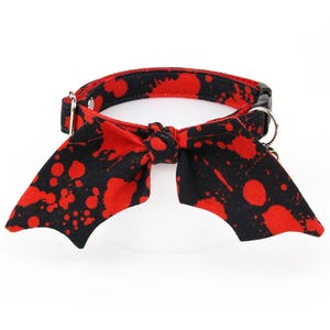 May include: A black pet collar featuring a red paint splatter pattern and a matching fabric bow tie shaped like bat wings. The collar includes a black plastic side-release buckle, a silver metal D-ring for a leash, and an adjustable metal slider.