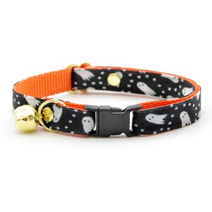 Peut inclure: Un collier de chat noir et orange avec un motif de fant&ocirc;mes blancs et une clochette dor&eacute;e. Le collier a une boucle en plastique noire.