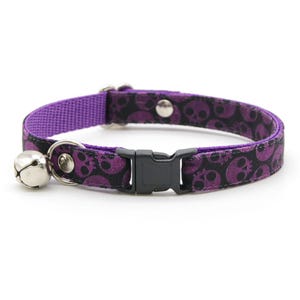 May include: Purple cat collar with a black skull pattern and a silver bell. The collar has a plastic buckle closure.