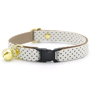 May include: A cat collar with a white fabric featuring black polka dots. The collar has a brown backing, a black plastic buckle, and a gold-colored bell. The collar is designed to be adjustable and is suitable for cats.