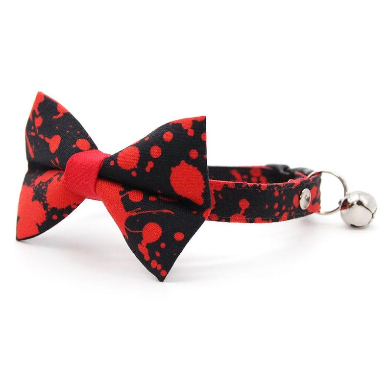 Op de afbeelding: Een halsband voor huisdieren met een zwarte stoffen basis en een rood verfspattenpatroon. De halsband is voorzien van een bijpassende stoffen strik aan de voorzijde en een kleine zilverkleurige metalen bel aan een ring aan de zijkant van de band.