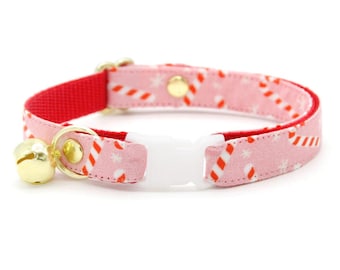 May include: A pink and red cat collar with a white plastic buckle and a gold bell. The collar features a pattern of red and white candy canes and snowflakes.