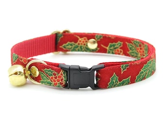 May include: A red cat collar with a festive holly and berry pattern. The collar features a gold-colored bell and buckle. The collar is approximately 0.5 inches wide and has a black plastic buckle.